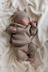 Een vredige pasgeboren baby gekleed in een gezellig, gebreid, 100% katoenen gebreid outfitje met een bijpassend gebreid broekje Cacao Cutie muts  