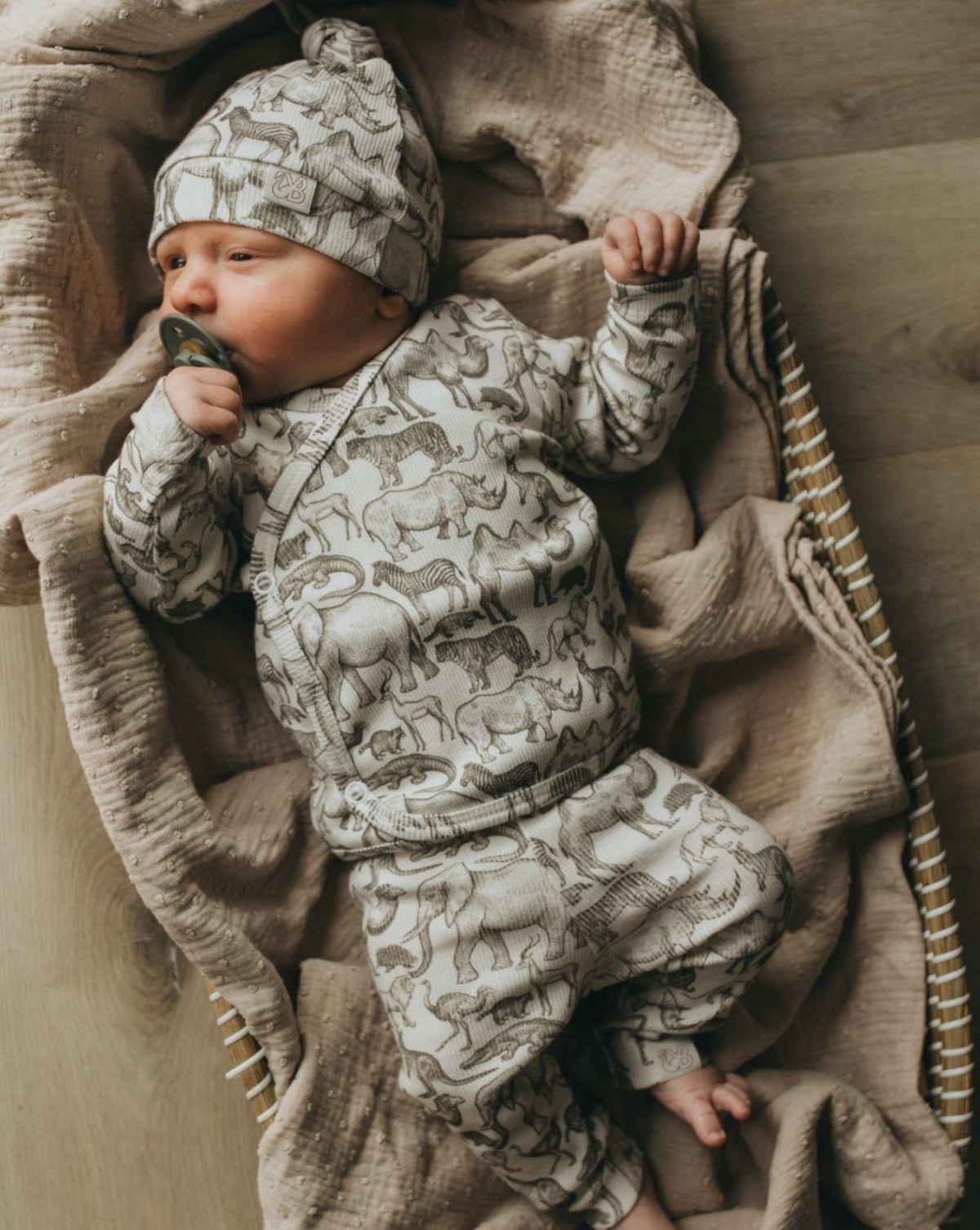 Een gezellige baby gekleed in een knus Yumi Baby Broekje Little Safari-outfit met een schattig bijpassend hoedje, ligt in een geweven mand, omringd door zachte, verkreukelde dekens, tevreden en vredig.