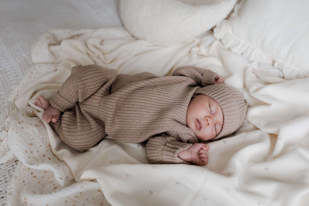 Een vredig kind gekleed in een knus Yumi Baby Knitted broekje Cacao Cutie-outfit met een bijpassende muts, slaapt comfortabel op een zacht, crèmekleurig textieloppervlak, met een zachte wieg van de hand