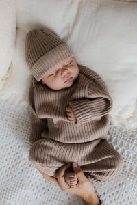 Een vredig kind gekleed in een knus Yumi Baby Knitted broekje Cacao Cutie-outfit met een bijpassende muts, slaapt comfortabel op een zacht, crèmekleurig textieloppervlak, met een zachte wieg van de hand