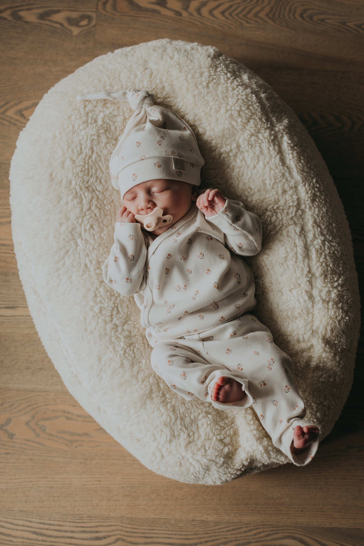 Een vredige pasgeboren baby die slaapt, gekleed in een schattig bedrukt Yumi Baby Broekje Peach Blossom onesie en bijpassend hoedje, liggend op een zacht wit oppervlak met de armen zachtjes omhoog bij het gezicht.