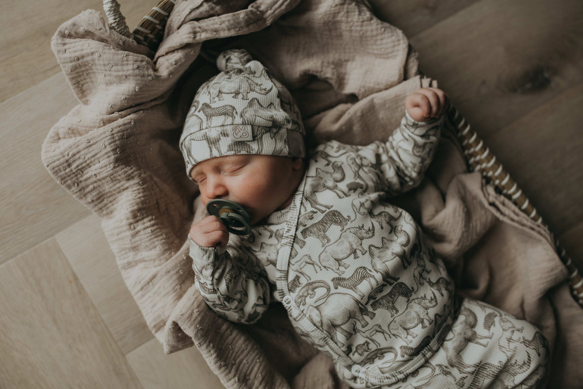 Een gezellig uitziende baby, gekleed in een Yumi Baby Overslagshirt Little Safari-onesie en bijpassend mutsje, rustend in een geweven mand op een zachte, milieuvriendelijke deken.