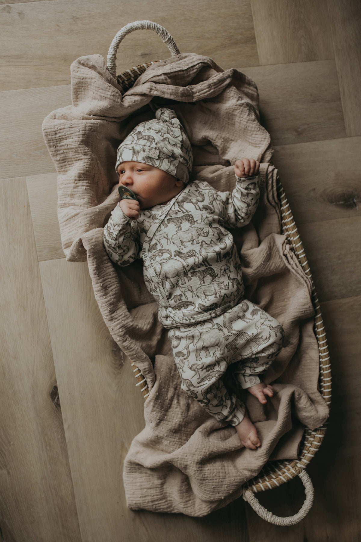 Een gezellig uitziende baby, gekleed in een Yumi Baby Overslagshirt Little Safari-onesie en bijpassend mutsje, rustend in een geweven mand op een zachte, milieuvriendelijke deken.