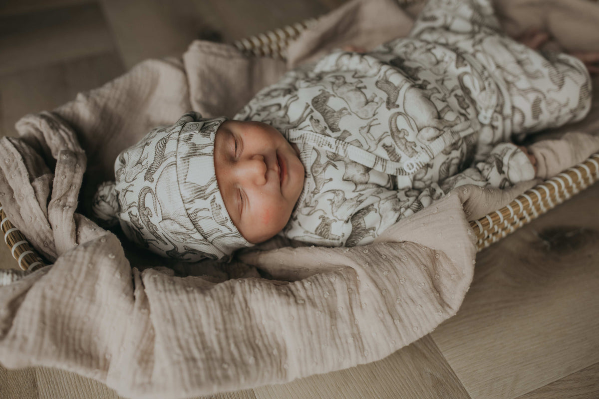 Een gezellig uitziende baby, gekleed in een Yumi Baby Overslagshirt Little Safari-onesie en bijpassend mutsje, rustend in een geweven mand op een zachte, milieuvriendelijke deken.