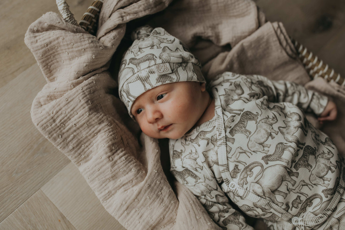 Een gezellig uitziende baby, gekleed in een Yumi Baby Overslagshirt Little Safari-onesie en bijpassend mutsje, rustend in een geweven mand op een zachte, milieuvriendelijke deken.