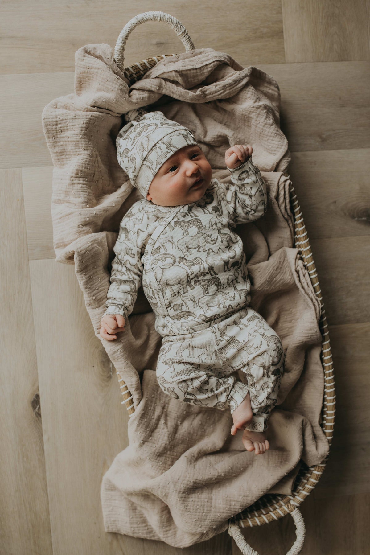 Een gezellig uitziende baby, gekleed in een Yumi Baby Overslagshirt Little Safari-onesie en bijpassend mutsje, rustend in een geweven mand op een zachte, milieuvriendelijke deken.