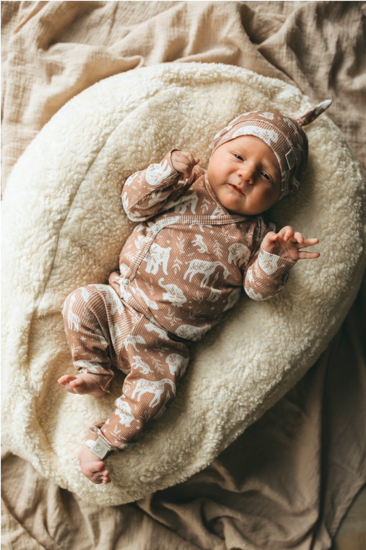 Een serene pasgeboren baby gekleed in een Yumi Baby Broekje Boho Jungle onesie gemaakt van OEKO-TEX gecertificeerd katoen en een bijpassend mutsje, vredig slapend in een knus rieten mandje gevoerd met een zachte, crèmekleurige deken.