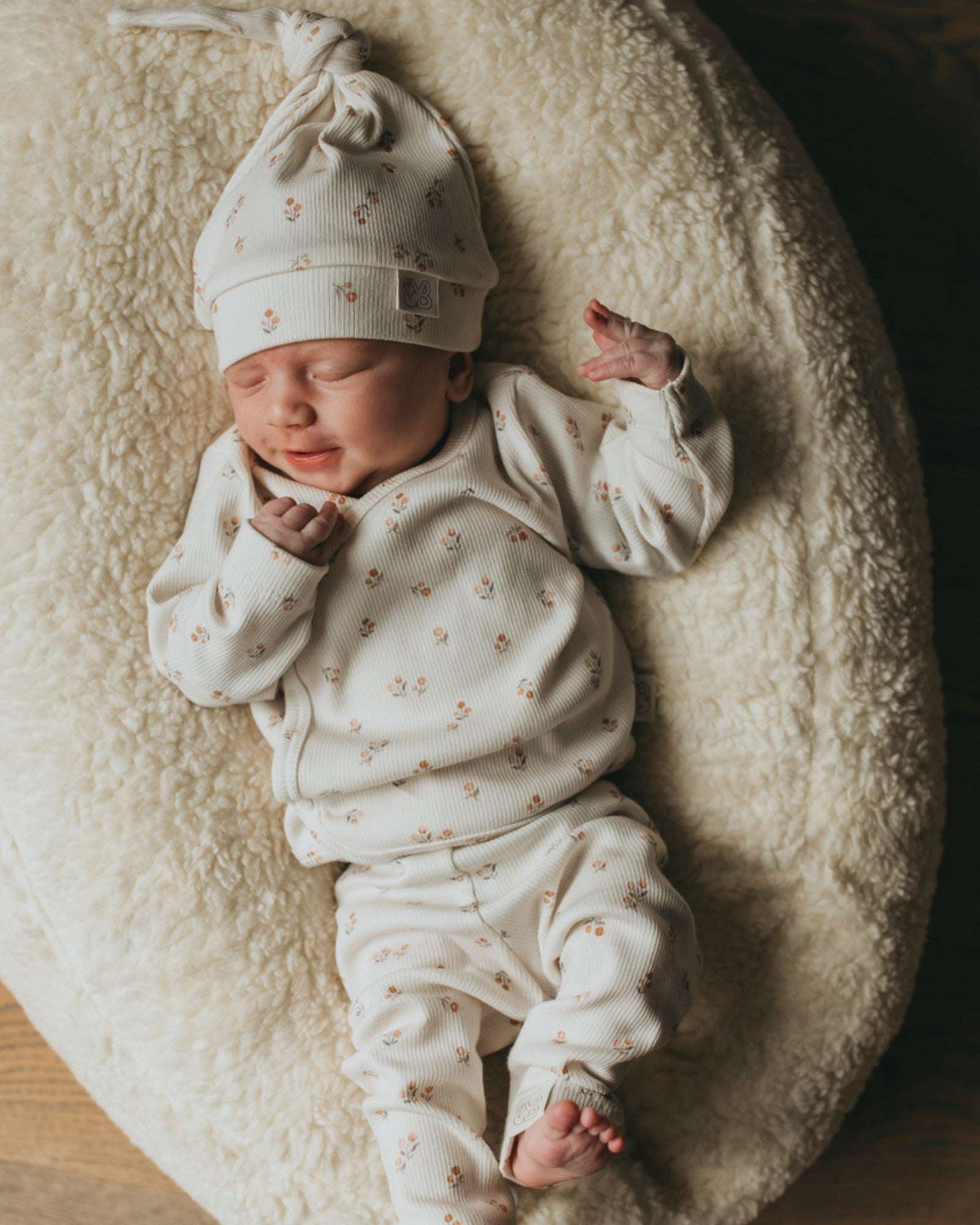 Een vredige pasgeboren baby die slaapt, gekleed in een schattig bedrukt Yumi Baby Broekje Peach Blossom onesie en bijpassend hoedje, liggend op een zacht wit oppervlak met de armen zachtjes omhoog bij het gezicht.