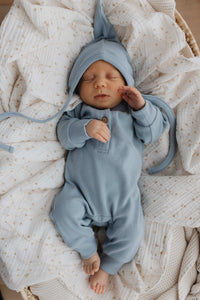 Een vredige pasgeboren baby slaapt heerlijk, gekleed in een Yumi Baby Boxpakje Blue Sky van hoogwaardig katoen met bijpassend mutsje, genesteld in een zacht crèmekleurig dekentje.