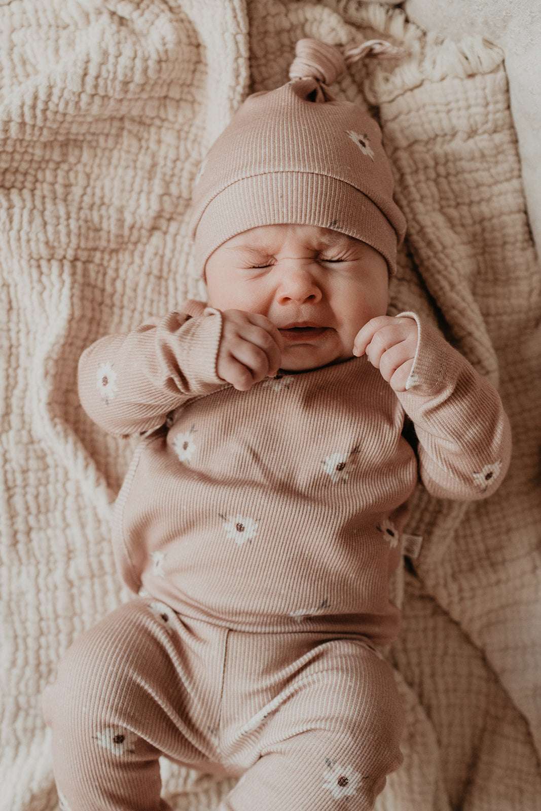 Een vredige pasgeboren baby, gekleed in een schattig, milieuvriendelijk broekje Zoë met stippen onesie met bloemenmotief en bijpassende muts, rekt zich een beetje uit of geeuwt, comfortabel liggend op een zachte, getextureerde Yumi Baby.