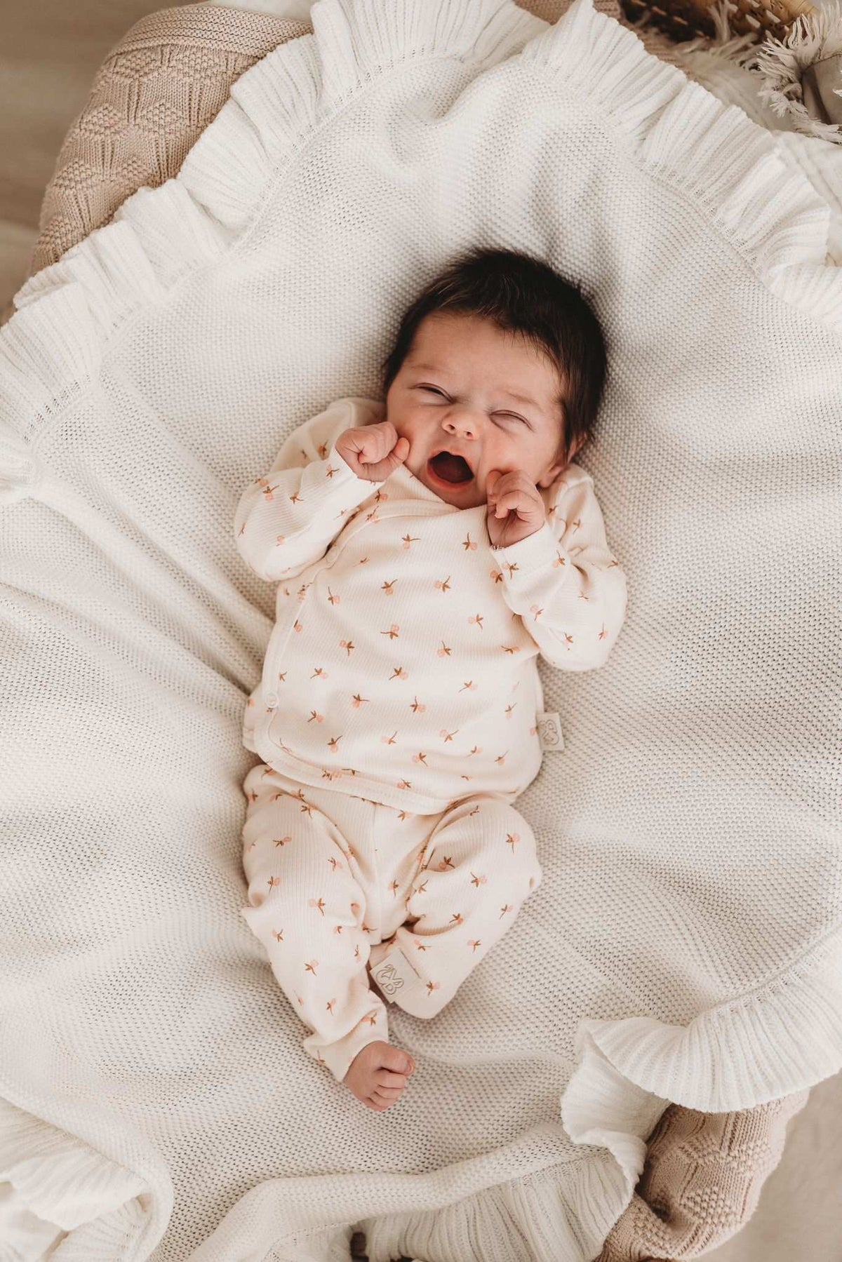 Een pasgeboren baby in een Broekje Tiny Tulip-outfit van Yumi Baby ligt op een crèmekleurige deken, geeuwend of huilend met kleine vuistjes bij het gezicht, en legt een kostbaar moment uit het vroege leven vast.