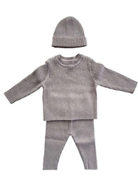 Een vredig kind gekleed in een knus Yumi Baby Knitted broekje Cacao Cutie-outfit met een bijpassende muts, slaapt comfortabel op een zacht, crèmekleurig textieloppervlak, met een zachte wieg van de hand