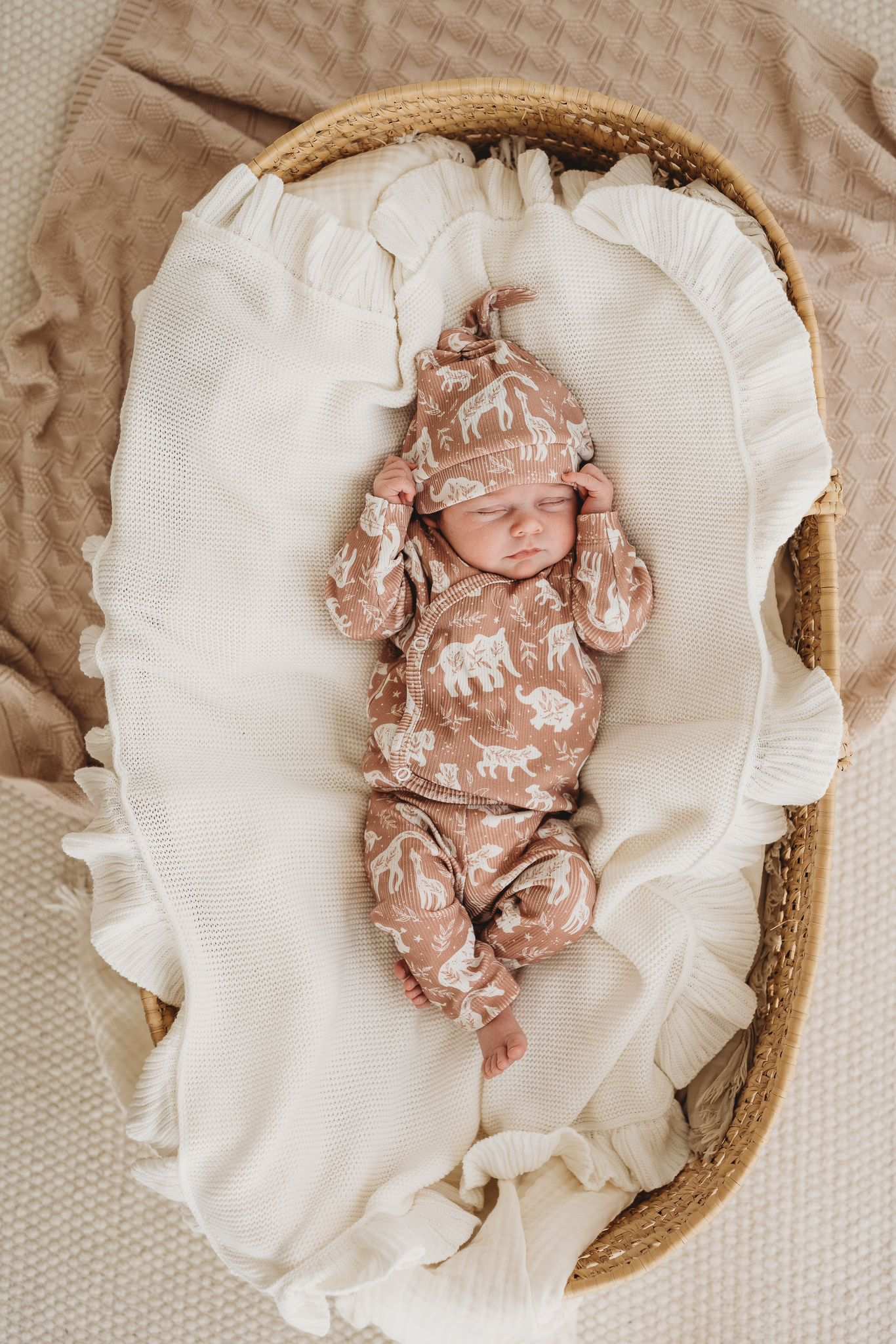 Een serene pasgeboren baby gekleed in een Yumi Baby Broekje Boho Jungle onesie gemaakt van OEKO-TEX gecertificeerd katoen en een bijpassend mutsje, vredig slapend in een knus rieten mandje gevoerd met een zachte, crèmekleurige deken.