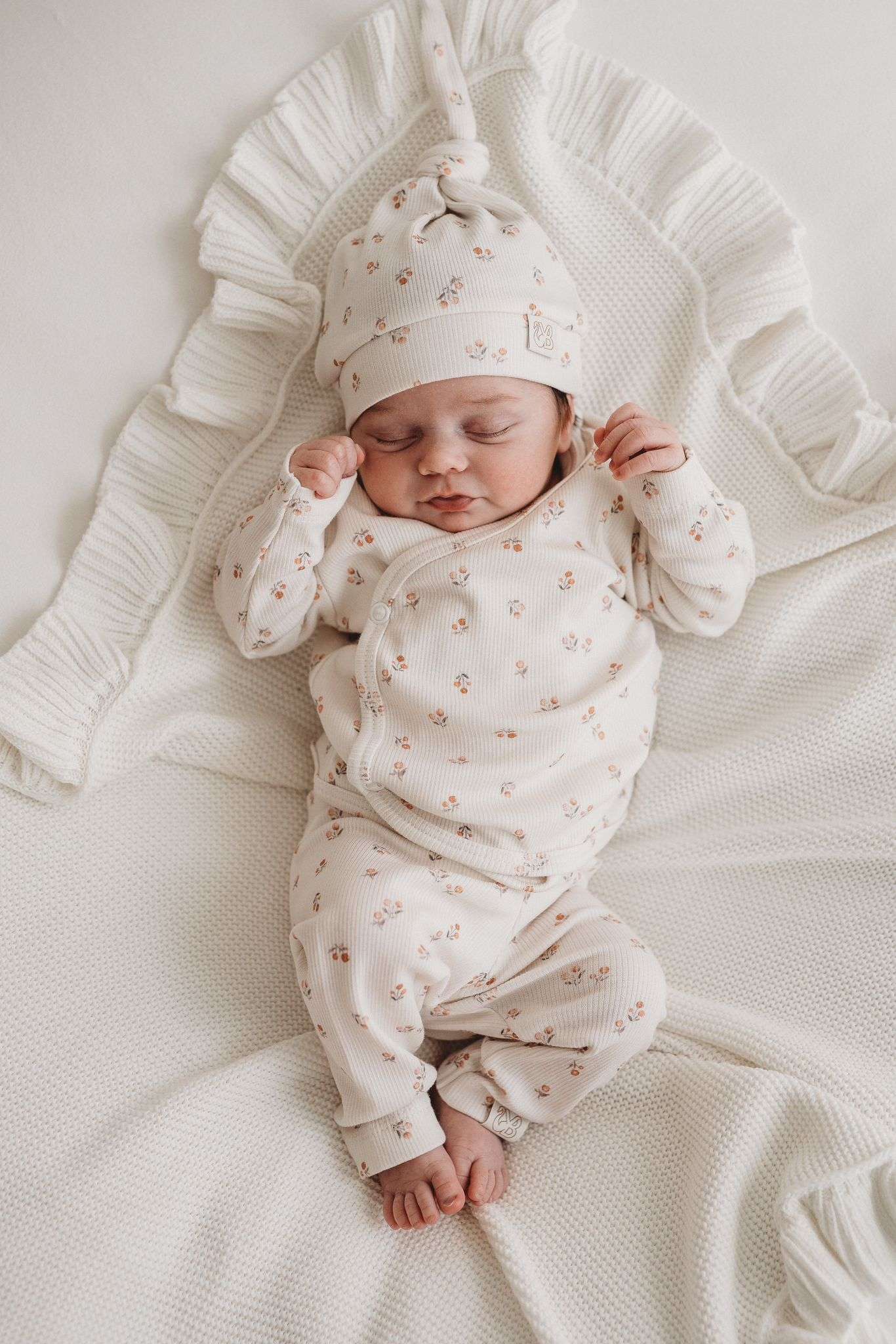 Een vredige pasgeboren baby die slaapt, gekleed in een schattig bedrukt Yumi Baby Broekje Peach Blossom onesie en bijpassend hoedje, liggend op een zacht wit oppervlak met de armen zachtjes omhoog bij het gezicht.