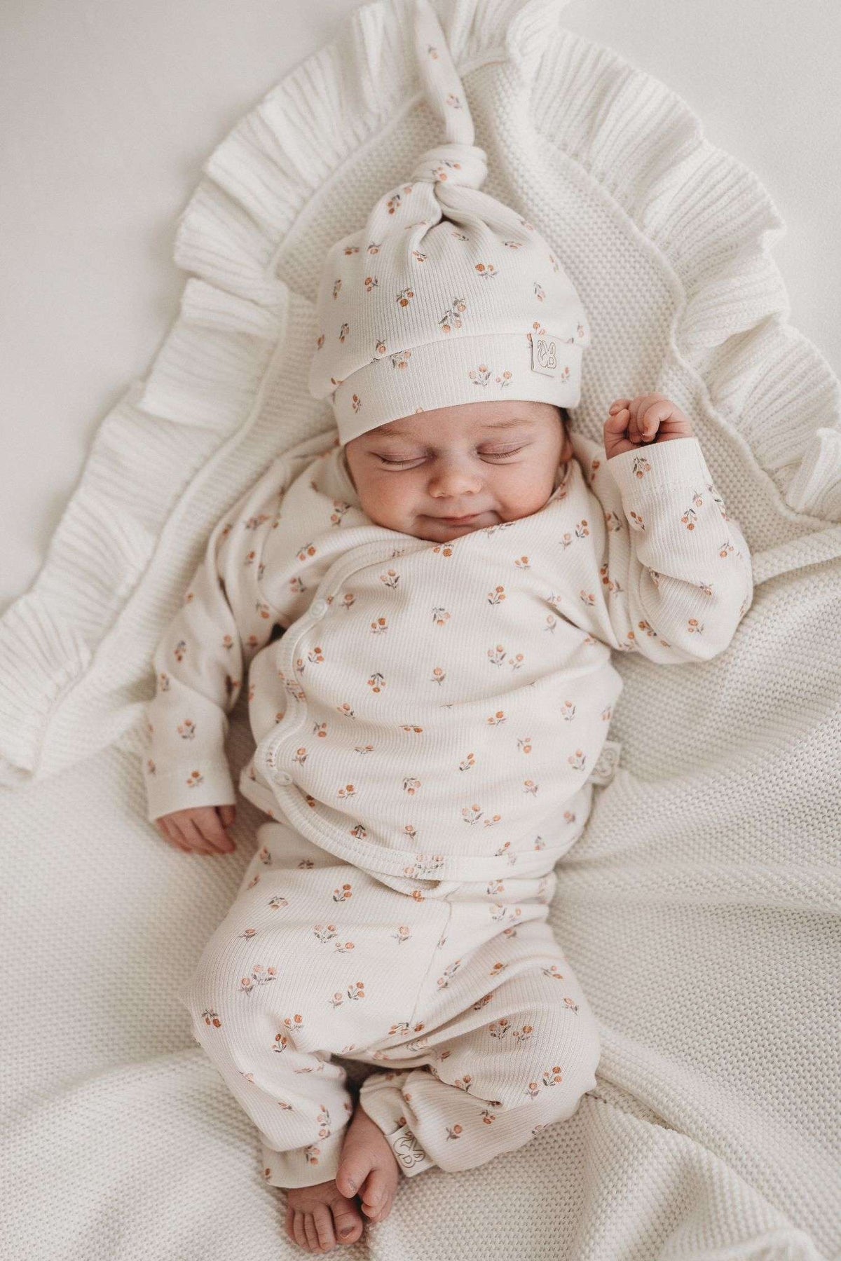 Een vredige pasgeboren baby die slaapt, gekleed in een schattig bedrukt Yumi Baby Broekje Peach Blossom onesie en bijpassend hoedje, liggend op een zacht wit oppervlak met de armen zachtjes omhoog bij het gezicht.