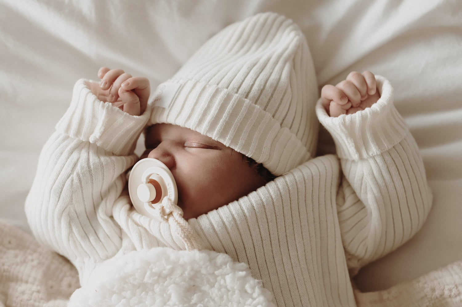 Een serene pasgeboren baby, gekleed in een Yumi Baby Gebreide Trui met Parelmoer, comfortabel genesteld in een geweven wiegje met een zachte deken, die vredige onschuld belichaamt.