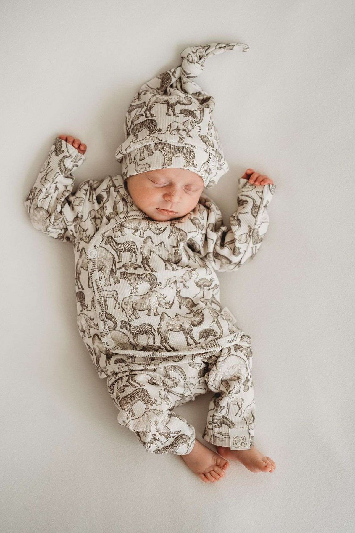 Een gezellig uitziende baby, gekleed in een Yumi Baby Overslagshirt Little Safari-onesie en bijpassend mutsje, rustend in een geweven mand op een zachte, milieuvriendelijke deken.